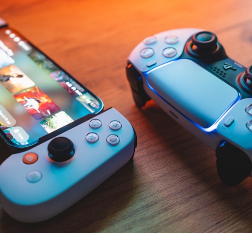 A gaming scenario featuring a smartphone with a game interface next to two controllers on a wooden surface. The smartphone displays vibrant images and icons, while the controllers feature a sleek, modern design with buttons and joysticks prominently visible. Warm lighting enhances the scene.